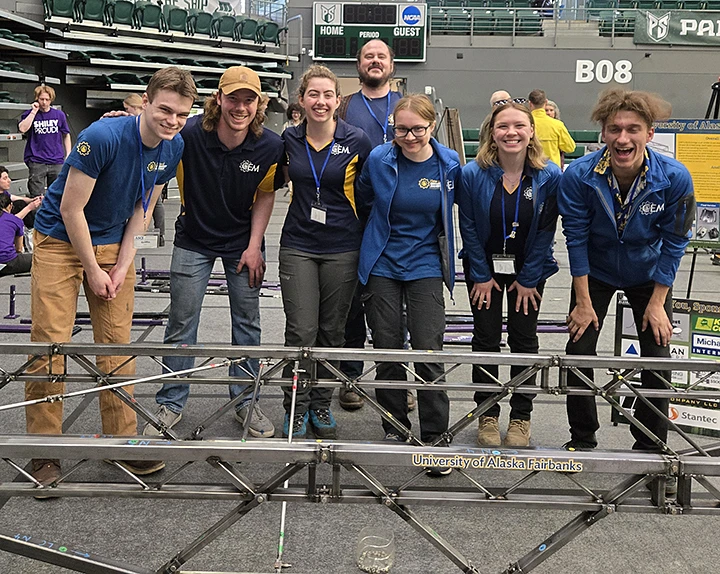 A group of seven people wearing matching team shirts pose together in an indoor event space beside a metal structure labeled "University of Alaska Fairbanks."
